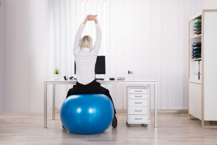 Rear View Of Businesswoman Sitting On Fitness Ball Stretching Her Arms In Office