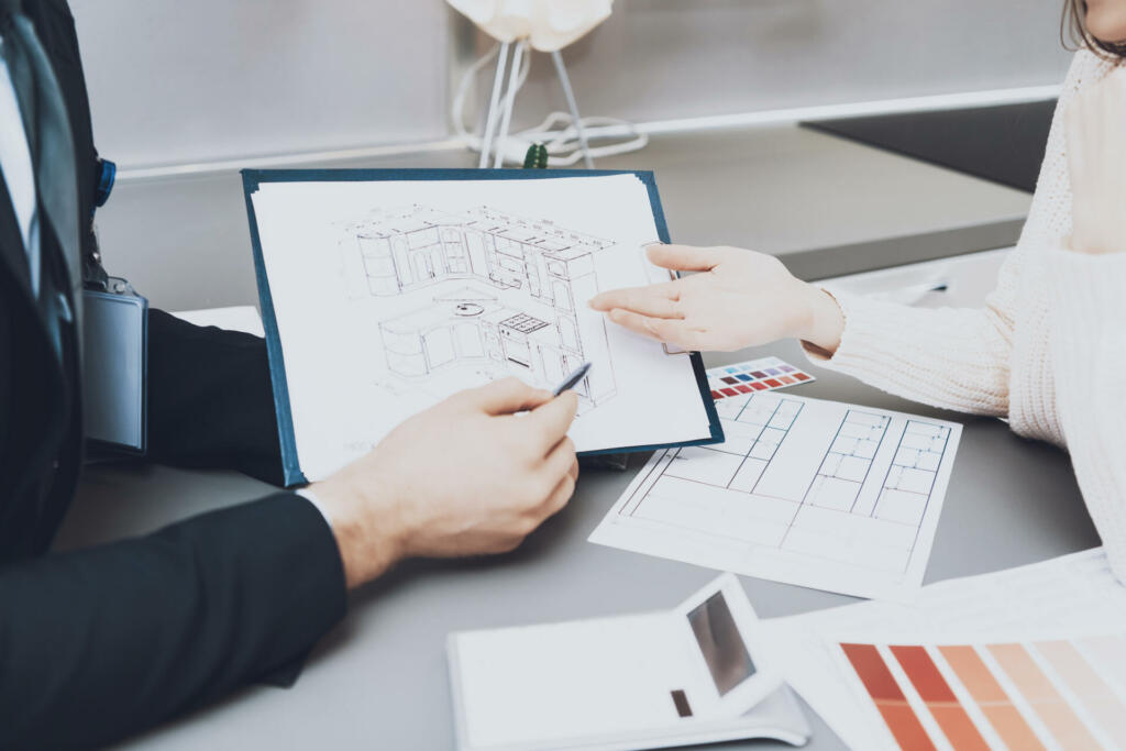 Close up manager in suit is showing blueprints to female client in kitchen store. The seller shows the kitchen layout and his idea in a design and flowers for her.