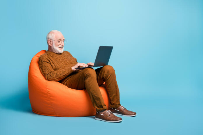 Portrait of his he nice, attractive focused bearded grey-haired man sitting in bag chair using laptop writing email letter browsing web wi-fi isolated over blue pastel color background