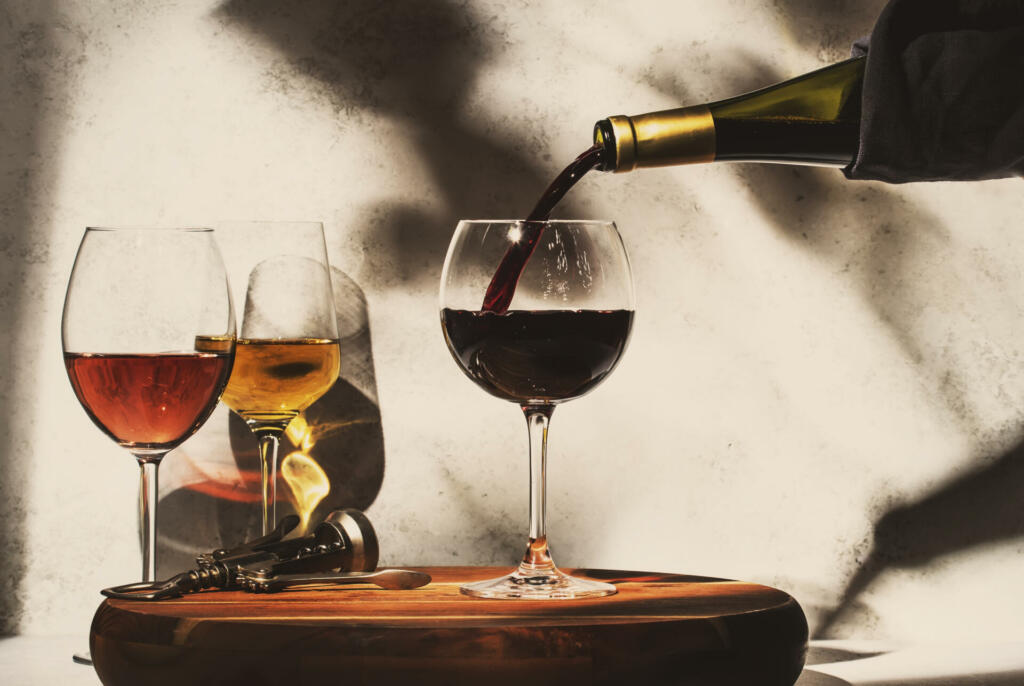 Wine tasting. Red wine pouring into glass on background with selection of red, white and rose wines in glasses. Hard sunlight and shadows from foliage