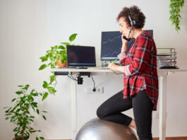 Woman teleworking at an adjustable standing desk with one knee resting on a fitball