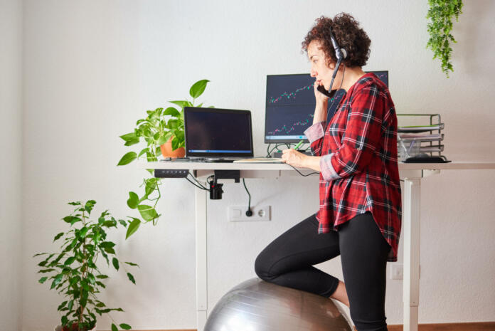 Woman teleworking at an adjustable standing desk with one knee resting on a fitball
