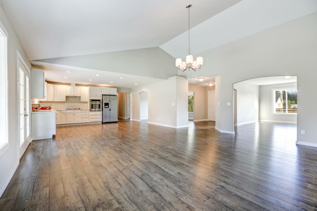 Spacious rambler home interior with vaulted ceiling over glossy laminate floor. Empty light filled dining or living space adjacent to new white kitchen room features pale grey walls. Northwest, USA