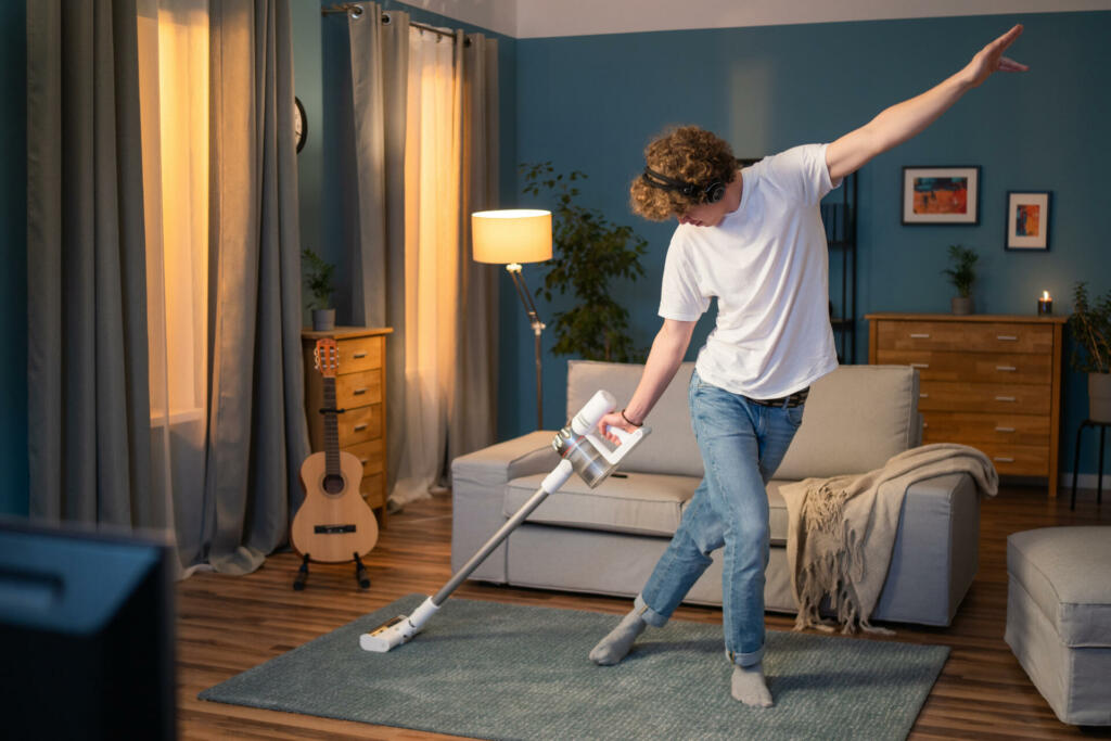 A boy is cleaning the living room in the evening. The man has fun vacuuming the carpet in the room, listening to music on wireless headphones and dancing with the vacuum cleaner.