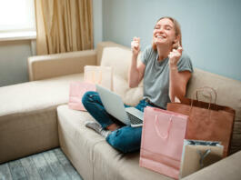 Online shopping at home. Young happy woman with is waiting for beggining of black Friday while sitting on a brown sofa with colorful shopping bags