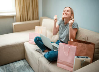 Online shopping at home. Young happy woman with is waiting for beggining of black Friday while sitting on a brown sofa with colorful shopping bags