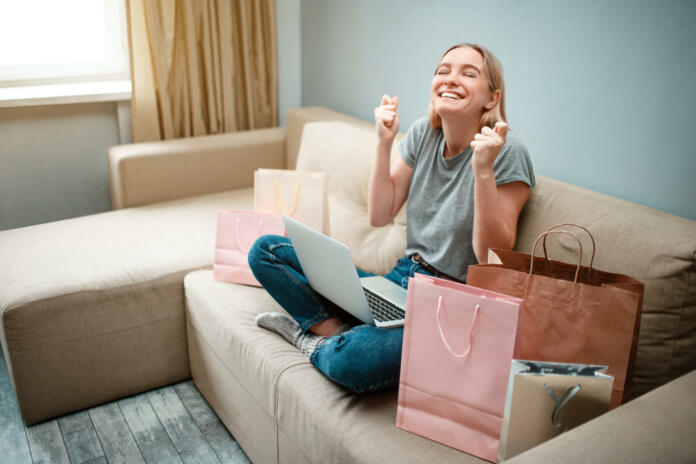 Online shopping at home. Young happy woman with is waiting for beggining of black Friday while sitting on a brown sofa with colorful shopping bags