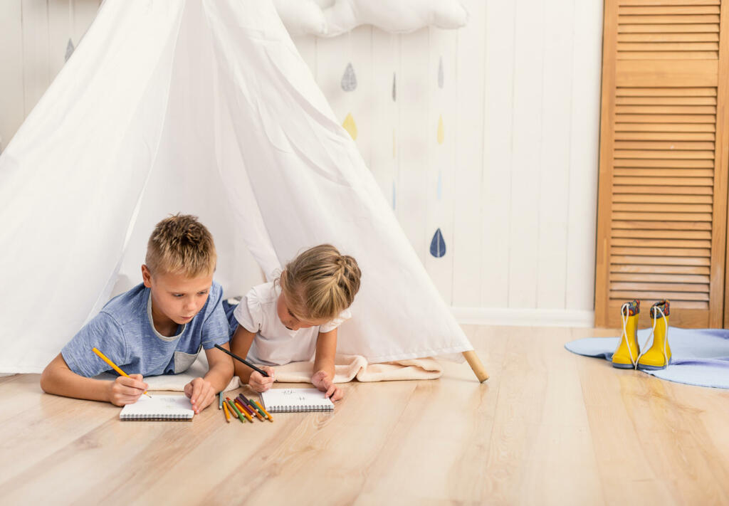 What are you drawing. Small cute brother and sister painting, using pencils while lying on floor in playroom