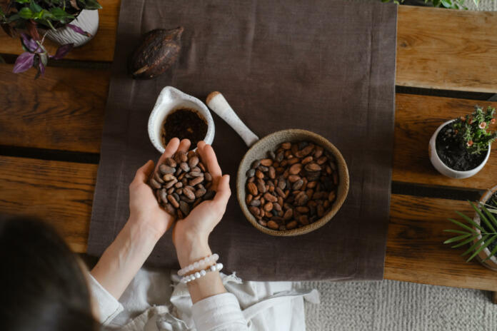 Ženske roke držijo kakavova semena, na mizi je skodelica vroče čoklade in posoda s kakavovimi semeni
