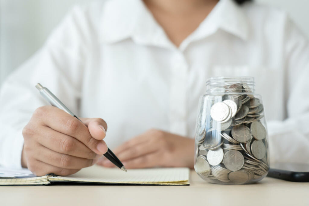 Woman with coins jar and checklist, expenses doing accounting plan. Writing down on a note book for planning.