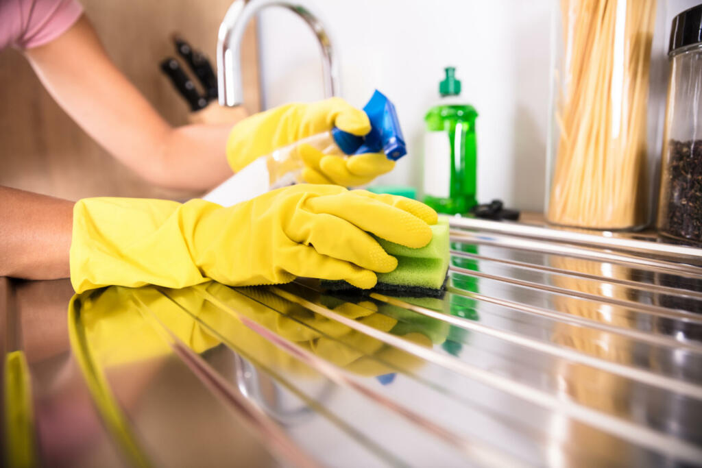 Close-up Of Person's Hand Wearing Yellow Gloves Cleaning Stainless Steel Sink With Sponge