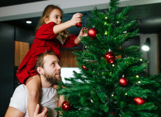 Izbrati naravno ali umetno smrečico? Cute little girl sitting on her dad's shoulders decorating a christmas tree with red shiny balls. Dad looking up at what she is doing.