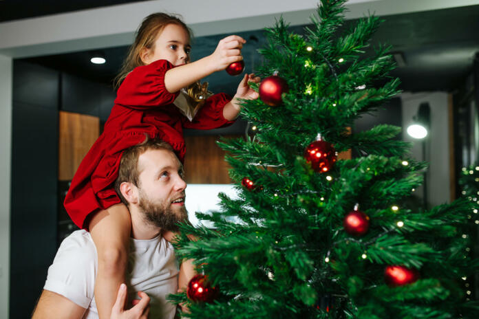 Cute little girl sitting on her dad's shoulders decorating a christmas tree with red shiny balls. Dad looking up at what she is doing.