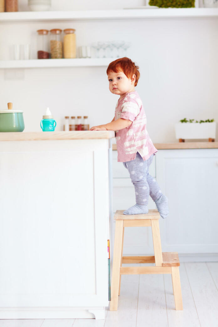 cute toddler baby climbs on step stool, trying to reach things on the high desk on the kitchen