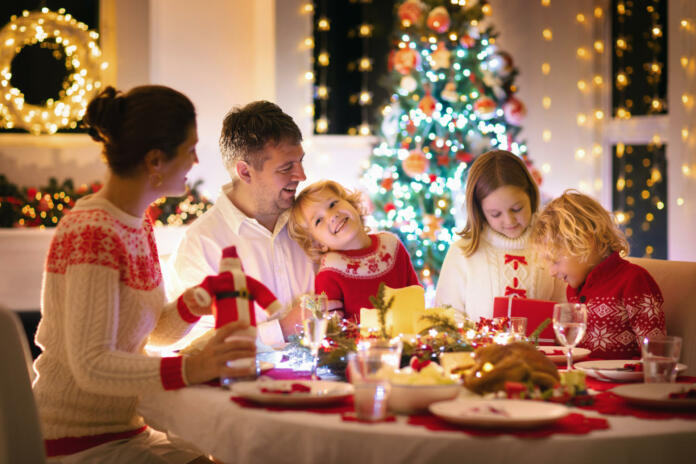 Family with children eating turkey Christmas dinner at fireplace and decorated Xmas tree. Parents, grandparents and kids at festive meal. Winter holidays celebration and food. Kids open presents.