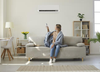 Skupinski nakup klime: Tako klimo kupite ceneje Woman who's sitting on sofa under warm plaid in living room switches off her air conditioner on wall. Young girl adjusting modern AC system, regulating temperature and enjoying cool fresh air at home