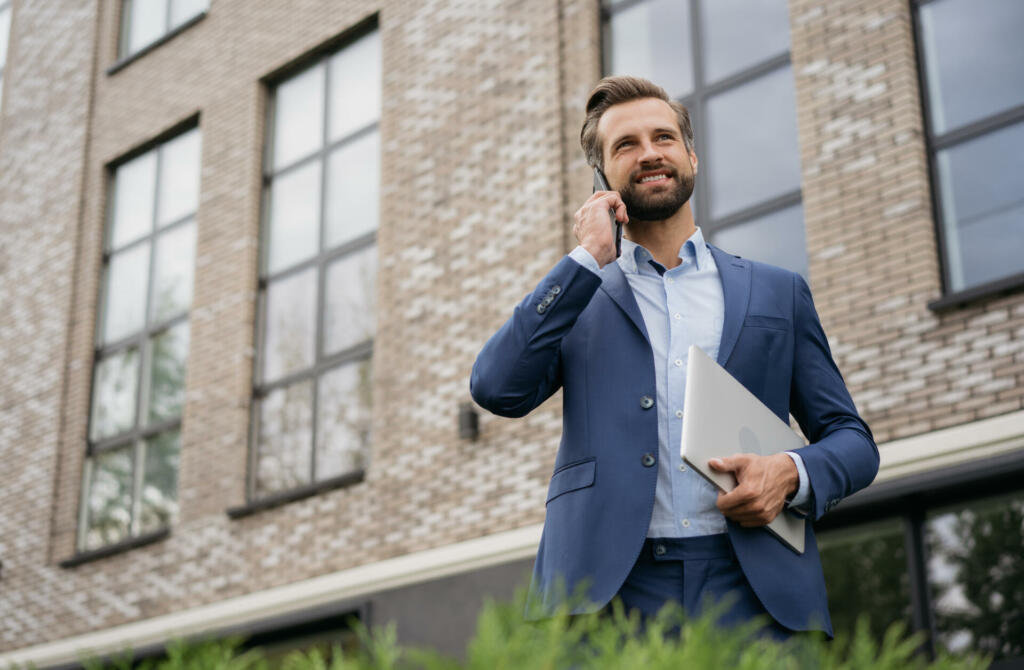 Handsome smiling businessman wearing stylish suit talking on mobile phone, holding laptop standing on the street. Successful business