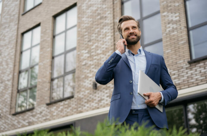 Handsome smiling businessman wearing stylish suit talking on mobile phone, holding laptop standing on the street. Successful business