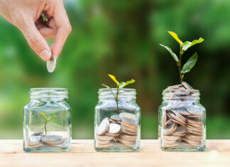 Varčevanje: Kako smiselna je metoda »The 5 Jar Method«? Money savings, investment, making money for future, financial wealth management concept. A man hand holding coin over stacked coins in glass jar and growing tree plant depicts Fund growth and wealth.