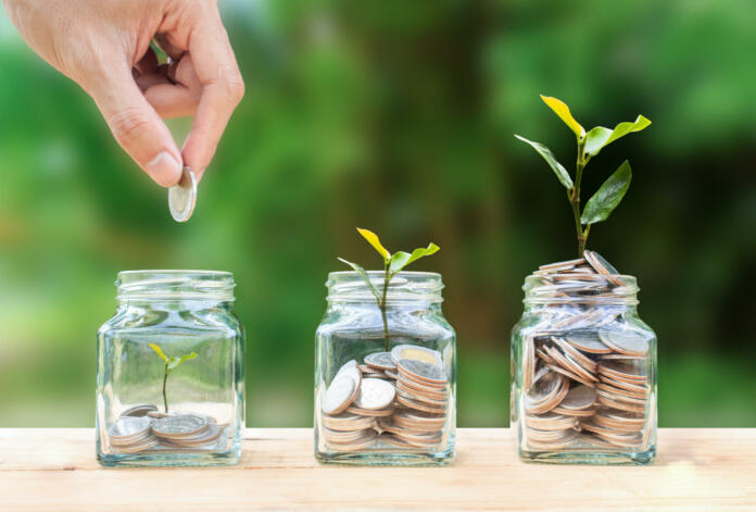 Money savings, investment, making money for future, financial wealth management concept. A man hand holding coin over stacked coins in glass jar and growing tree plant depicts Fund growth and wealth.