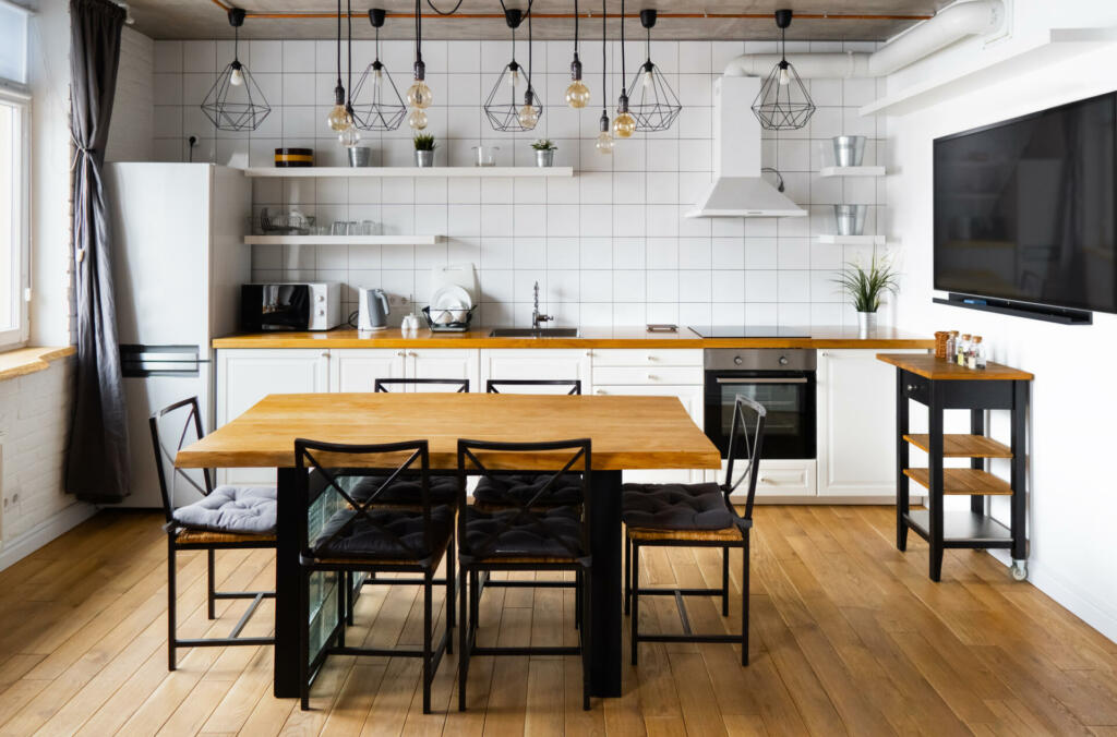 An eat-in kitchen interior design in modern scandinavian style with big wooden table and chairs against light wood floor, bright white walls and furnitures with TV, appliances and hanging light bulbs