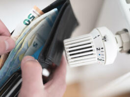 close-up view of person holding wallet with cash next to turned down thermostat on radiator, rising energy and heating costs concept