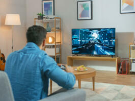 In the Living Room Man Sitting on a Couch Holds Controller Playing in a Console Video Game, 3D Action Shooter Gameplay Shown on TV Screen.