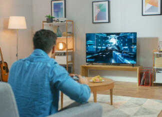 Kako daleč naj bo televizija glede na velikost zaslona? In the Living Room Man Sitting on a Couch Holds Controller Playing in a Console Video Game, 3D Action Shooter Gameplay Shown on TV Screen.