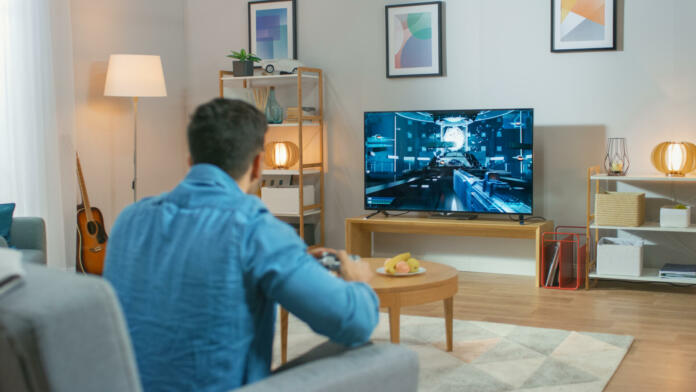 In the Living Room Man Sitting on a Couch Holds Controller Playing in a Console Video Game, 3D Action Shooter Gameplay Shown on TV Screen.