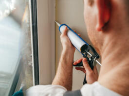Young man wearing overalls sealing cracks between window and trim using waterproof silicone caulk on the balcony.
