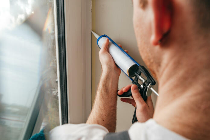 Young man wearing overalls sealing cracks between window and trim using waterproof silicone caulk on the balcony.