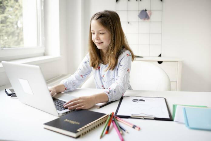Portrait of happy little european blond girl pupil in study online using laptop at home, smiling small child, homeschooling concept