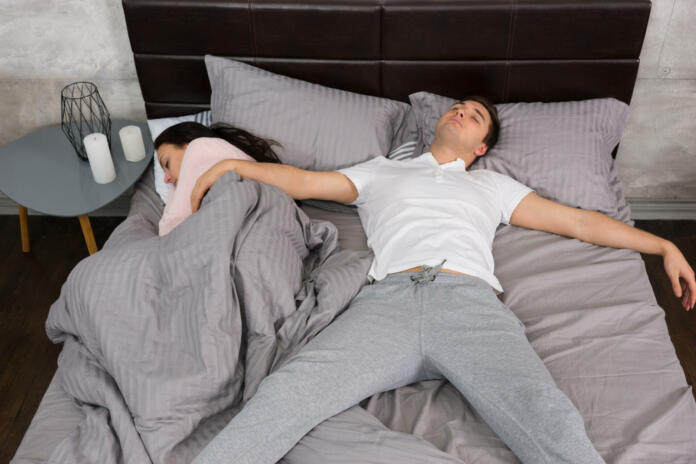 Young male sleeping in free fall position with his girlfriend occupied the whole bed, wearing pajamas, near bedside table with candles