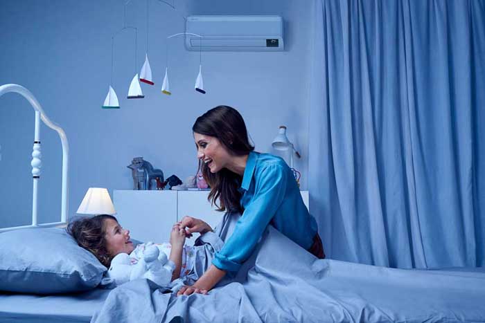 Mama pokriva hčerko v postelji, na steni je klimatska naprava