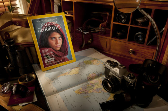 Alston, UK - July 2, 2011: A classic edition of the National Geographic magazine circa. 1985, lies on a wooden bureau surrounded by a selection of typical photo-journalist equipment including a Pentax K1000 35mm film camera, a Billingham 335 camera bag, a Philips Great World Atlas and several Optomax and Asahi camera lenses.