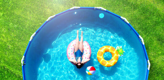 Aerial. Girl resting in a metal frame pool with inflatable toys. Summer leisure and fun concept. Frame pool stand on a green grass lawn. Top view from drone.
