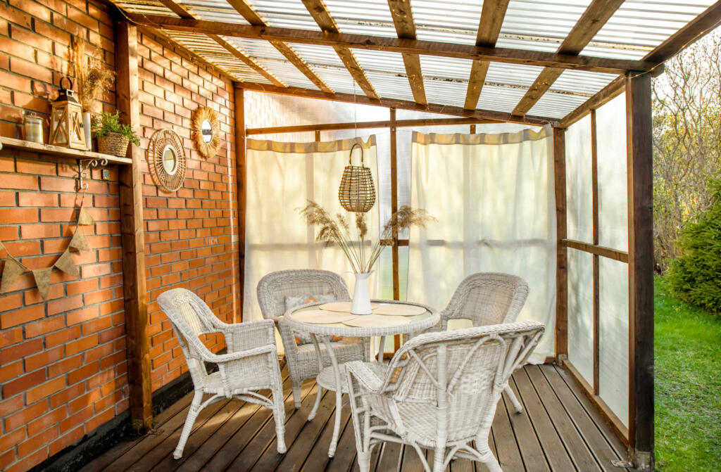 Boho style home patio terrace with curtains and wicker, rattan, reed decorations. Wall made with greenhouse plastic and ceiling is polycarbonate. White garden furniture, mirrors red on brick wall.