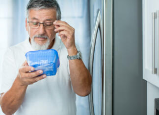 Mit ali resnica? Hrana s pretečenim rokom je še vedno užitna? Is this still fine? Senior man in his kitchen by the fridge, looking at the expiry date of a product she took from her fridge -