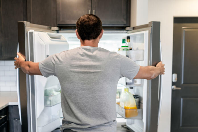 Hrbtna stran lačnega človeka, ki odpira vrata hladilnika, gospodinjskih aparatov in išče hrano v sodobni kuhinji.