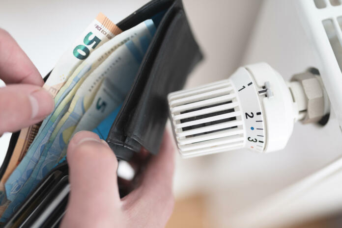 close-up view of person holding wallet with cash next to turned down thermostat on radiator, rising energy and heating costs concept