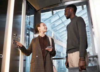 Koliko virusa COVID-19 izdihne oboleli? Za okužbo je dovolj vožnja z dvigalom Low angle portrait of two young coworkers chatting in glass elevator at modern office building