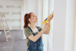 Young woman applying glue from a gun to a window frame during home renovations in an unfinished living room