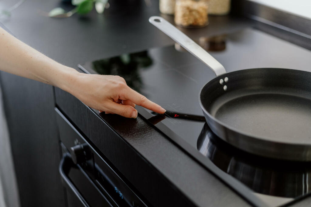 female hand touching sensor button on control panel of electrical hob and cooking dinner on frying pan at home kitchen, modern household appliance