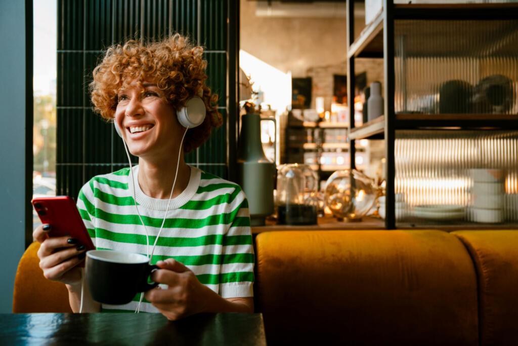 Young ginger curly woman in headphones using cellphone while drinking coffee in cafe