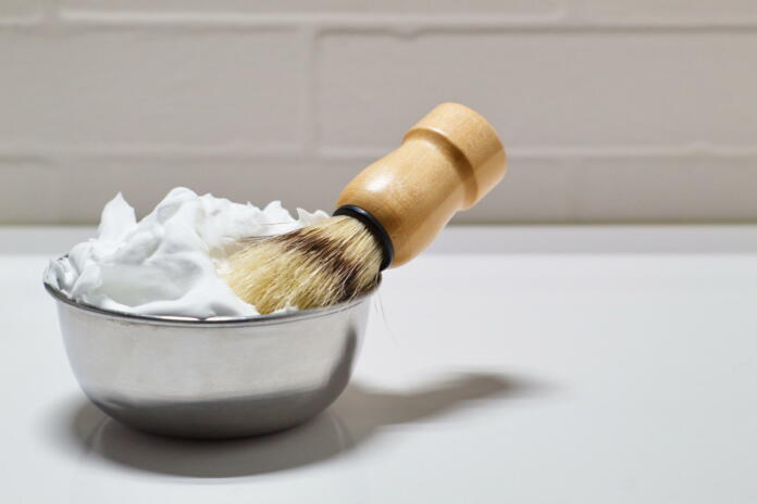 Bowl with shaving foam and brush on white background in bathroom for daily grooming