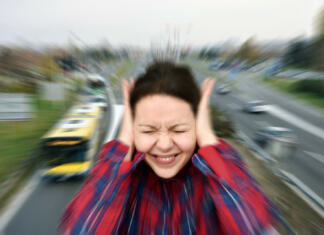 Razkrivamo: Kako hrup vpliva na zdravje in kakšne so rešitve? Woman with closed eyes covering her ears in the street while vehicles are passing by fast in the background. Stressful and frustrating situation. Noise pollution concept. Radial blur filter applied.