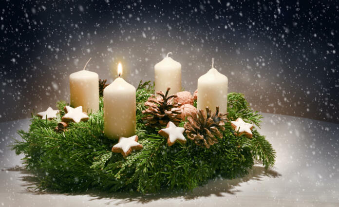 Advent wreath from evergreen branches with white candles, the first is burning for the time before Christmas, dark snowy background with copy space, selected focus