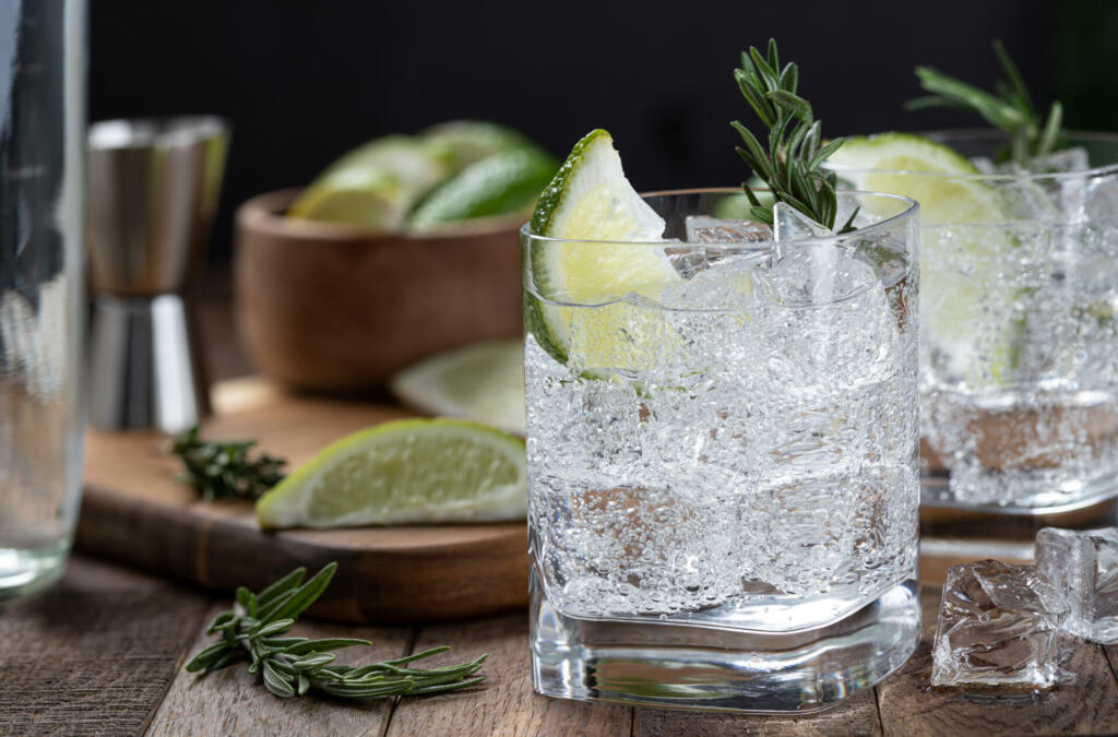Gin and tonic cocktail with lime slices, rosemary and ice on a rustic wooden table