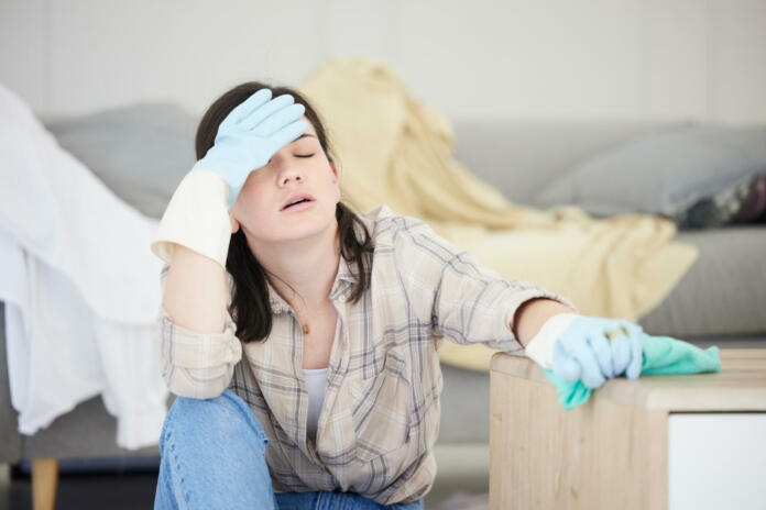 Tired, stressed young woman with headache doing spring cleaning chores, sitting in her living room at home. Frustrated, overworked female is exhausted from hygienic domestic housekeeping service