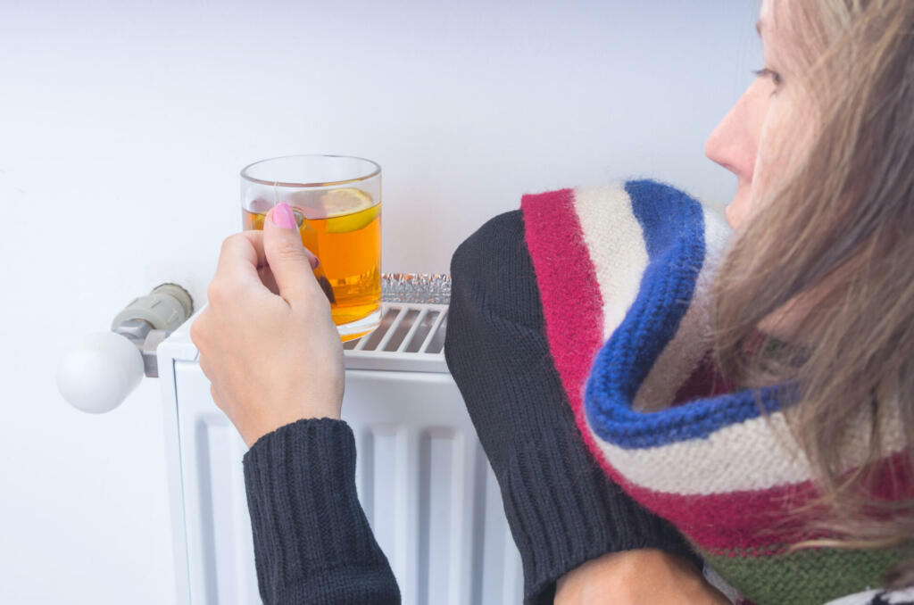 A cup of tea with lemon in the hands of a young woman, basking near a heating radiator at home in winter. The symbolic image of cold weather, colds, home heating. Selective focus.